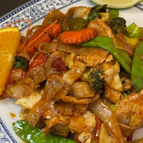 A close-up of a colorful stir-fry dish with noodles, vegetables, and chicken, garnished with orange and lime.